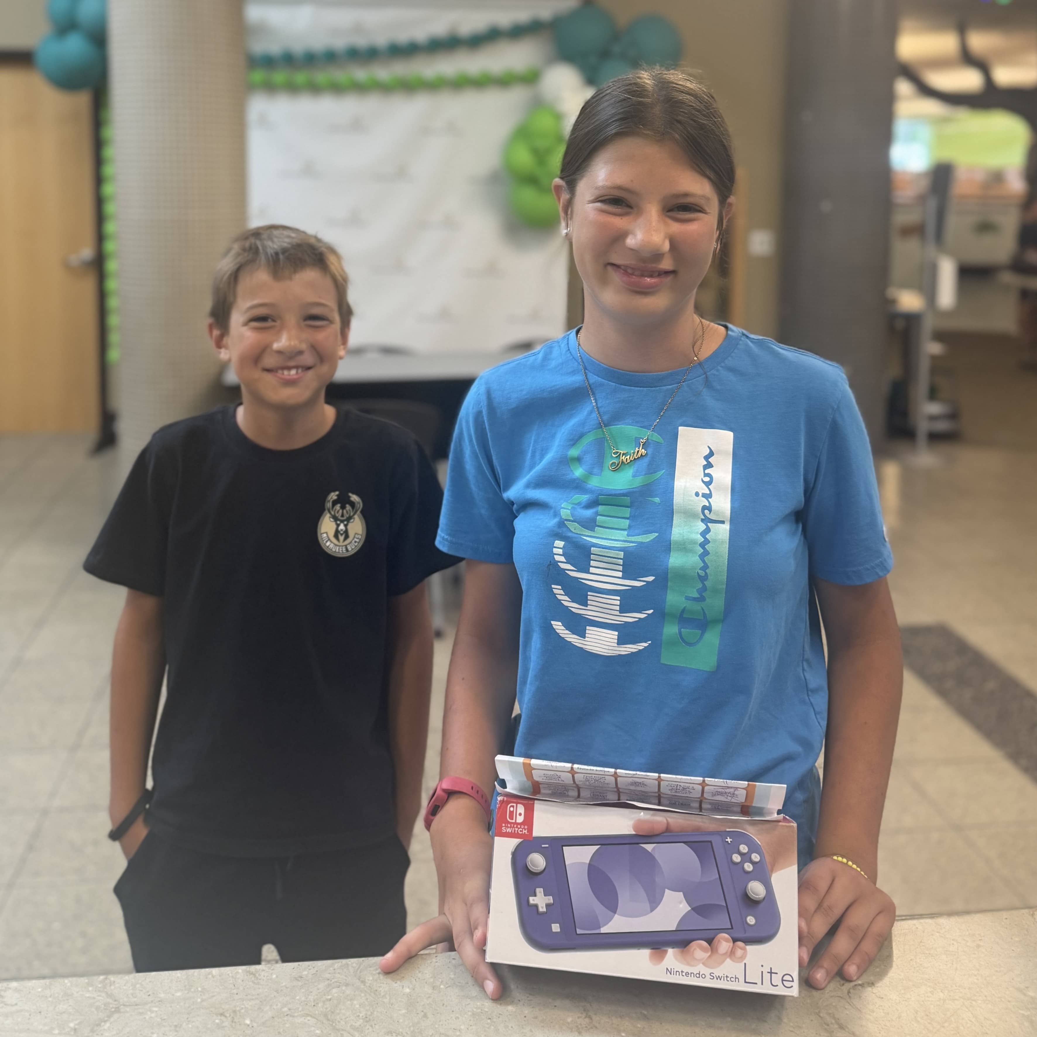 A smiling girl holding a Nintendo Switch as her younger brother smiles from behind
