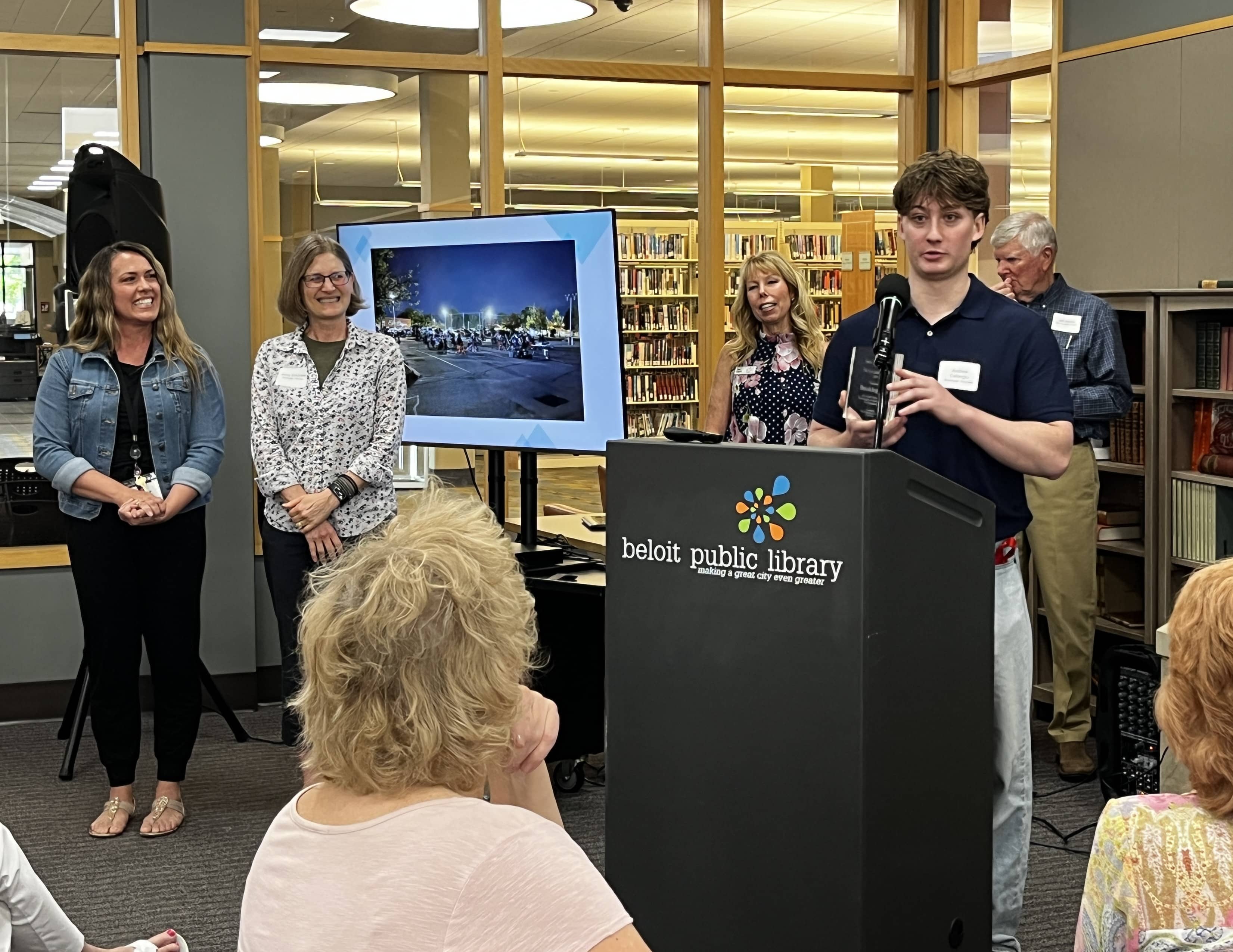Young man in dark polo speaking at a podium while four others stand behind smiling