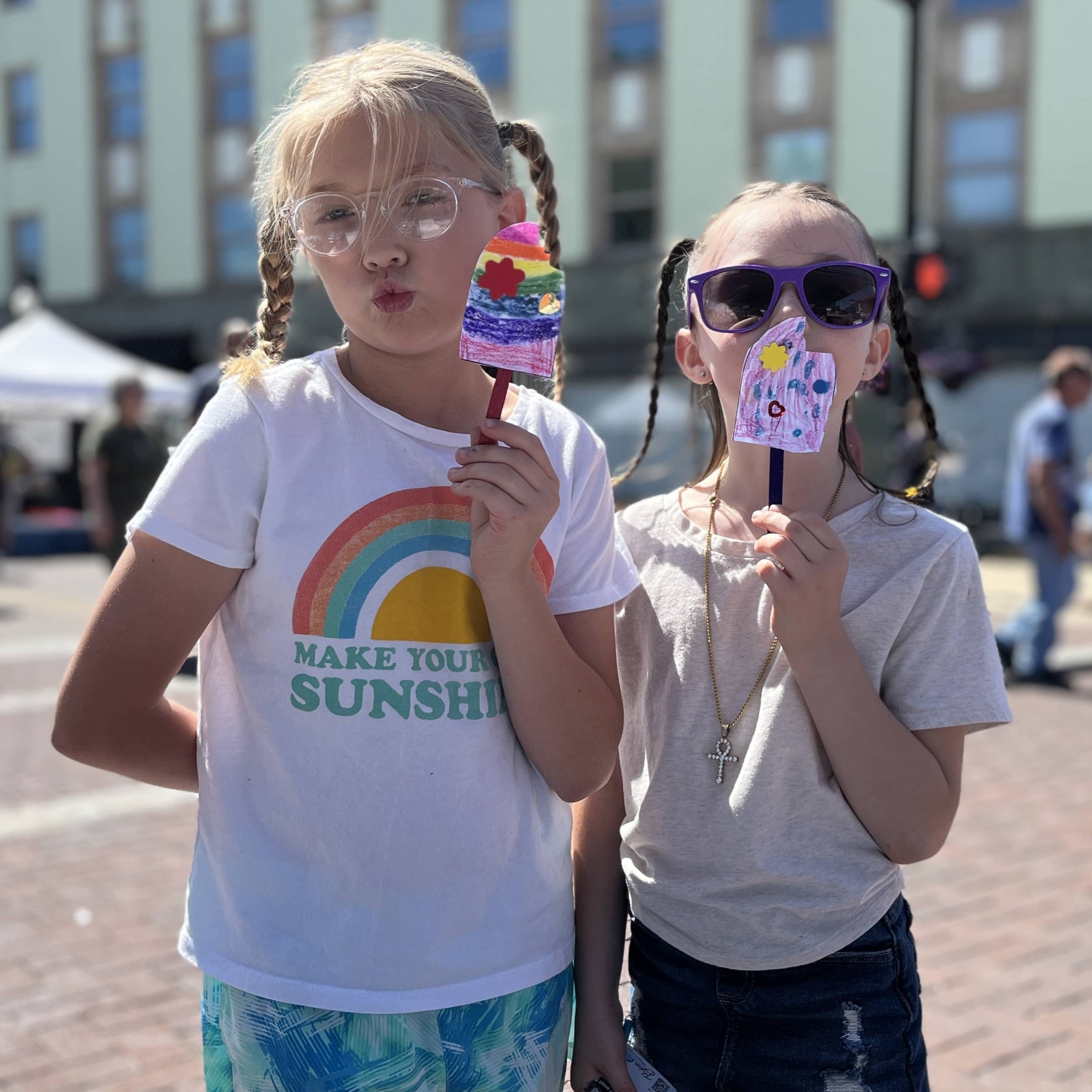 Two girls posing in sunny downtown Beloit with popsicle crafts they made at the Farmers Market