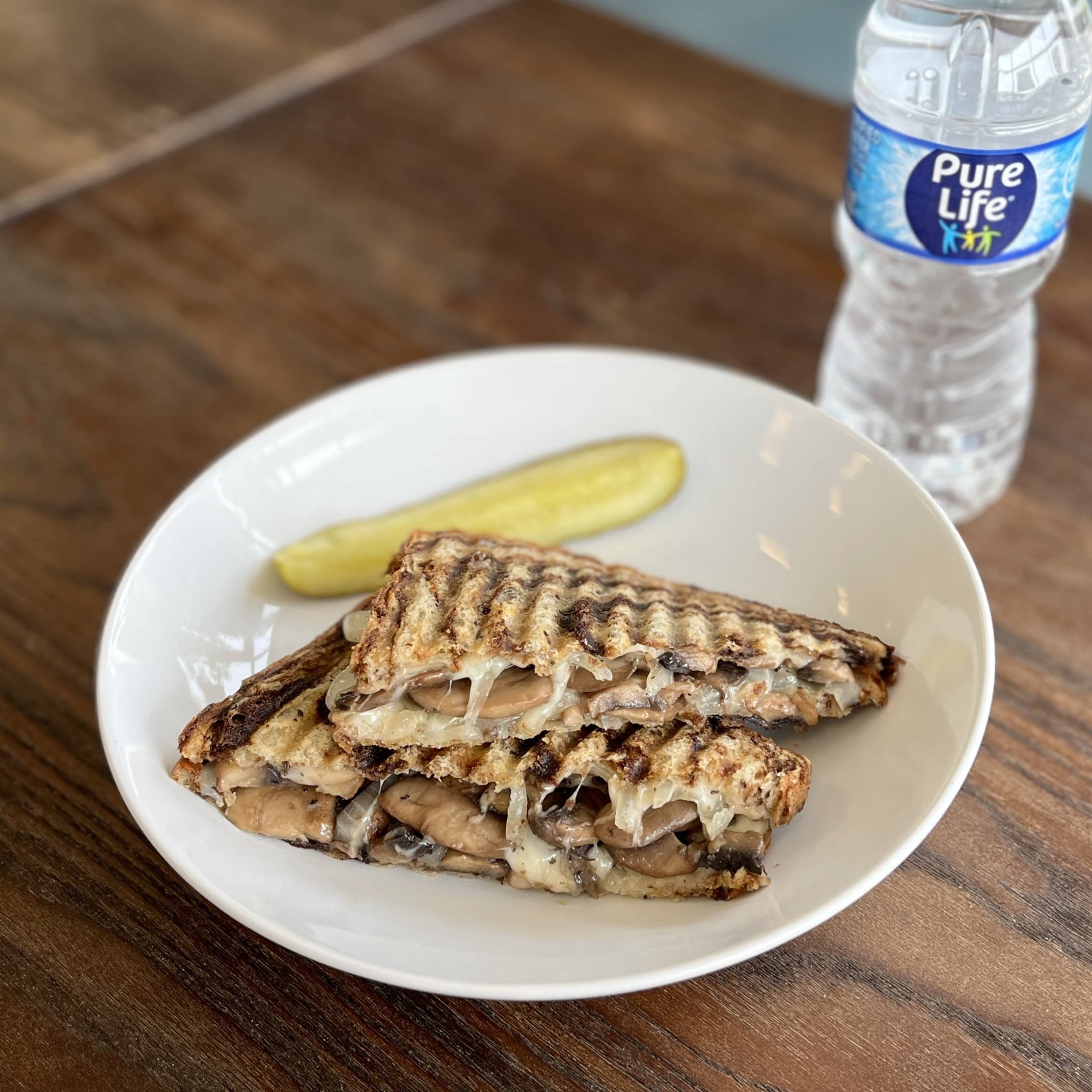 A white plate on a table with a sandwich cut in half and pickle with a water bottle next to it