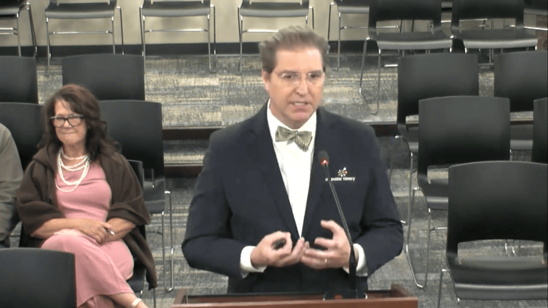 Man in suitcoat and bowtie gesturing and speaking in front of a podium