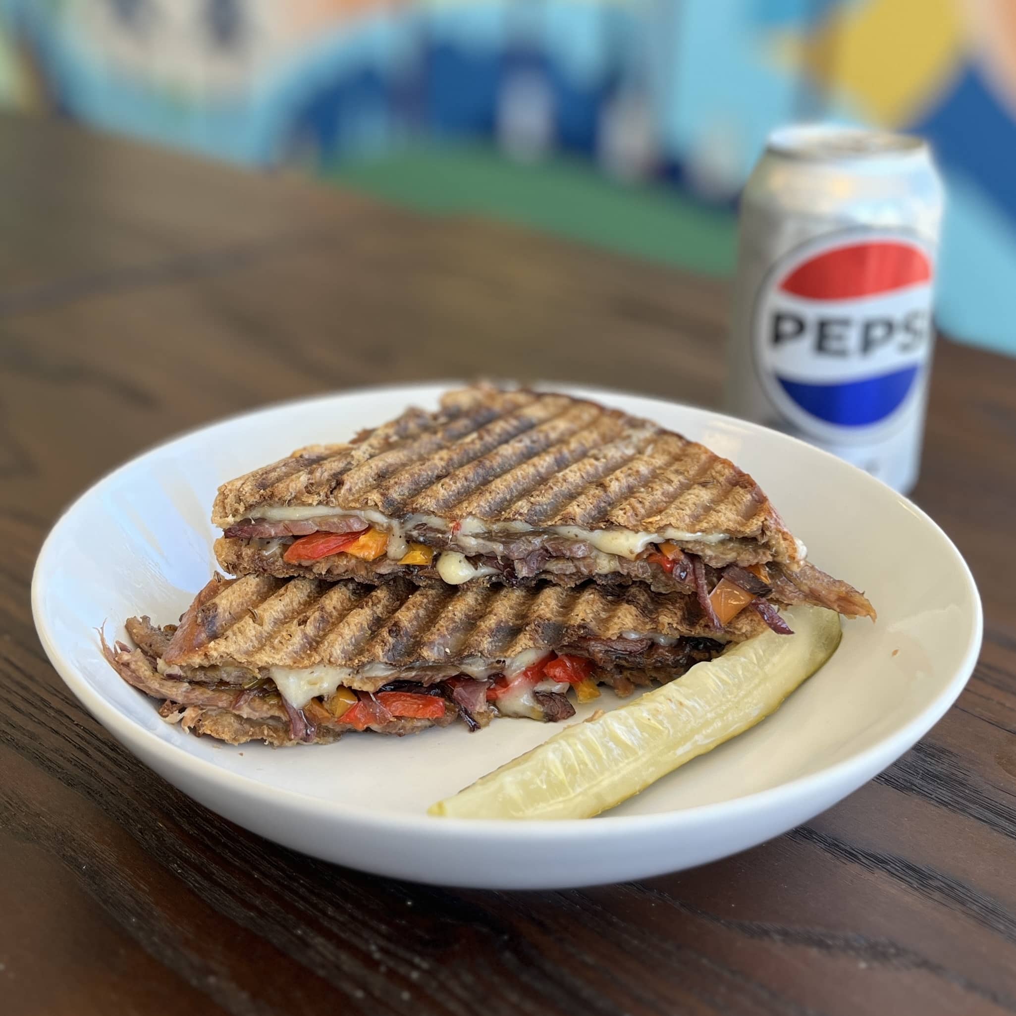 A white plate on a table with a sandwich cut in half and pickle with a water bottle next to it