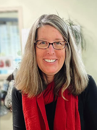 Person with light brown hair, glasses, a black top, and a red scarf smiling at the camera. The background is softly blurred with greenery.