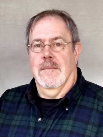Middle-aged man with glasses and a plaid shirt against a gray background.